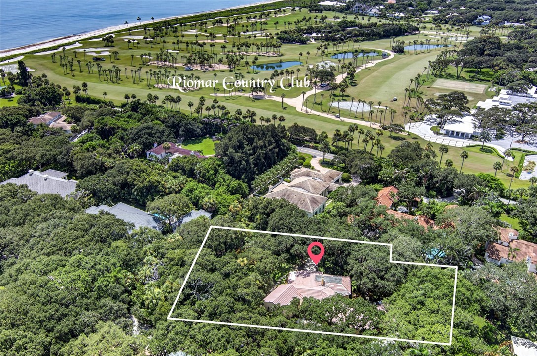 Aerial perspective of a property amidst dense trees, featuring a light-colored roof and a prominent red location marker