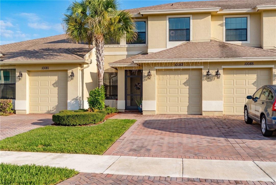 Stucco exterior residence with a tiled roof, featuring a paver driveway, an attached garage, and a palm tree landscaping element