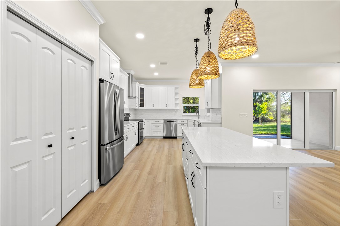 Spacious kitchen featuring a large island with a light-toned countertop, wood-finish flooring, white cabinetry, stainless steel appliances, and woven pendant lighting