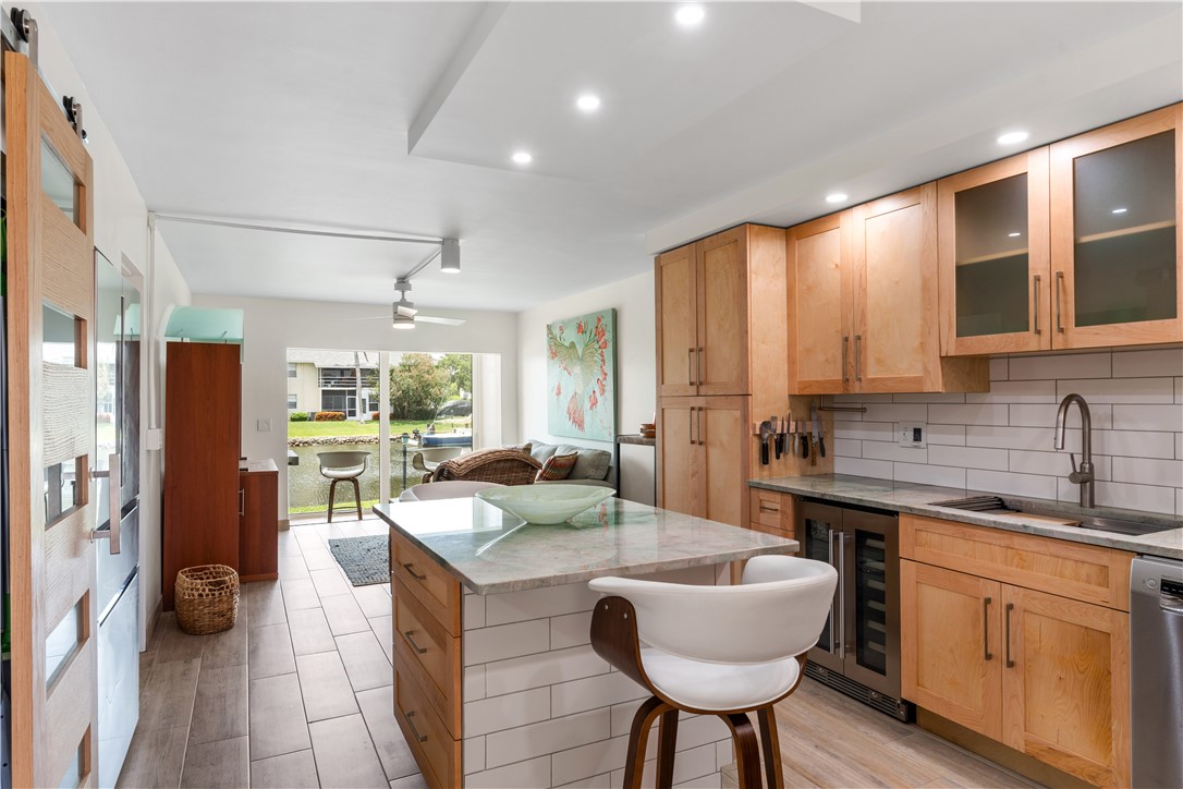 Open-concept kitchen featuring light wood cabinetry, stone countertops, a tile backsplash, and stainless steel appliances, including a built-in wine cooler