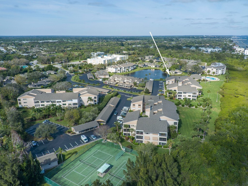 Multi-story residential buildings set within a landscaped community featuring a tennis court, parking areas, and a community pool