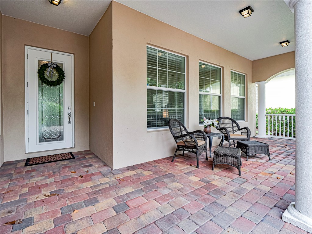 Covered front patio featuring a multi-toned paver surface, decorative column, and architectural windows