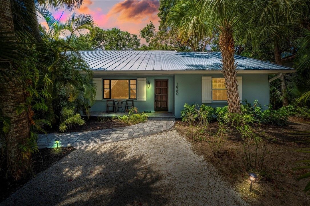 Coastal-style home with a metal roof and light blue exterior, featuring a wood front door, decorative white window shutters, and a paver pathway