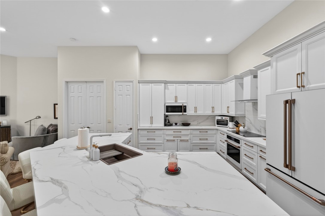 Spacious kitchen featuring a large island with an undermount sink, white cabinetry with bronze-finish hardware, integrated appliances, and light-toned countertops with veining