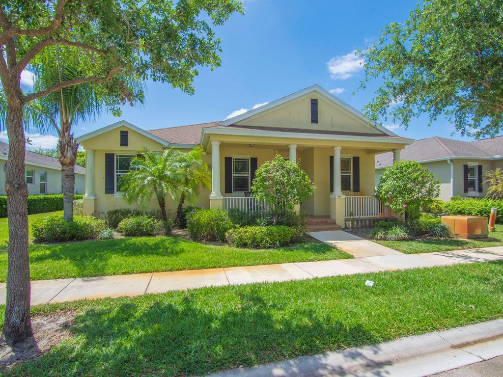 The property features a well-maintained lawn and mature trees, a covered front porch with white pillars, and a light-colored exterior with dark window shutters