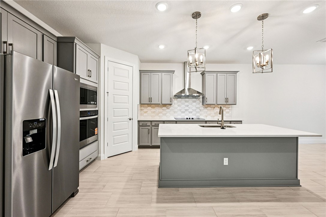 Kitchen featuring gray cabinetry, appliances with stainless steel finishes, pendant lighting, a kitchen island with sink, and light stone countertops