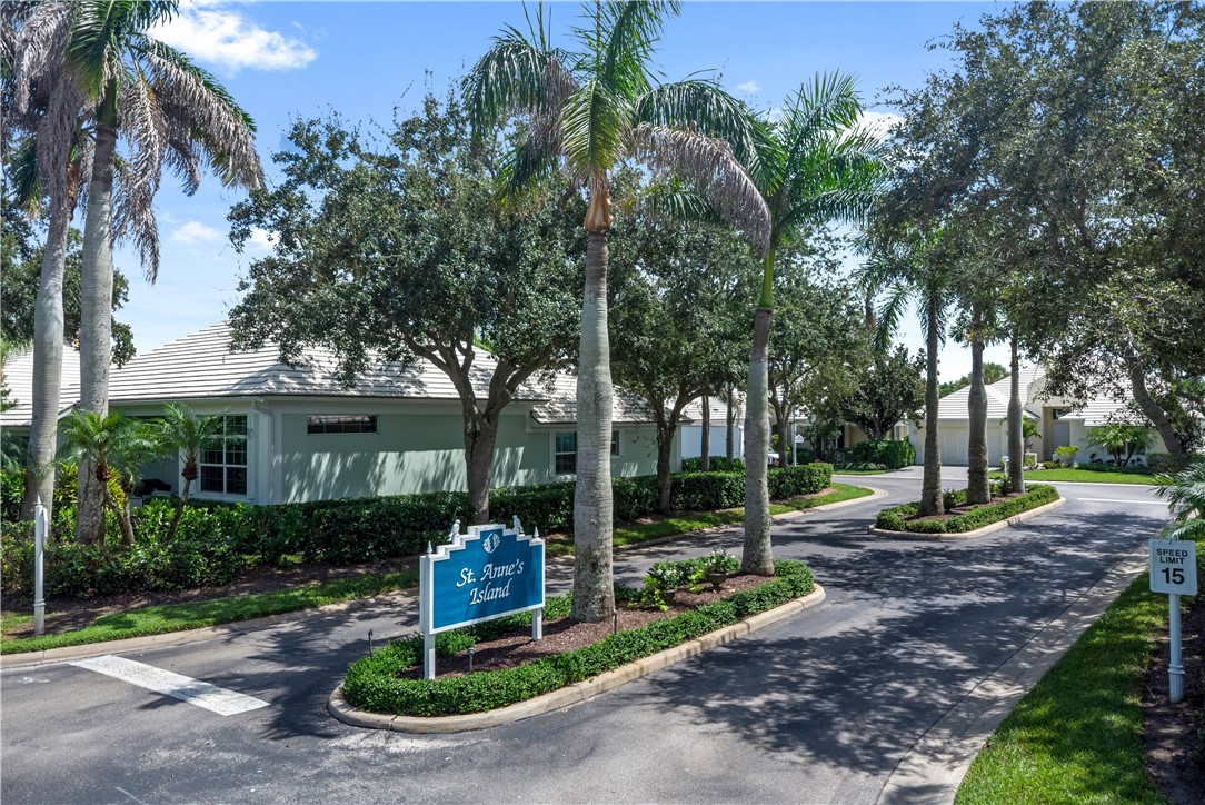 Paved entrance with mature palm trees, manicured shrubbery, and a visible community sign