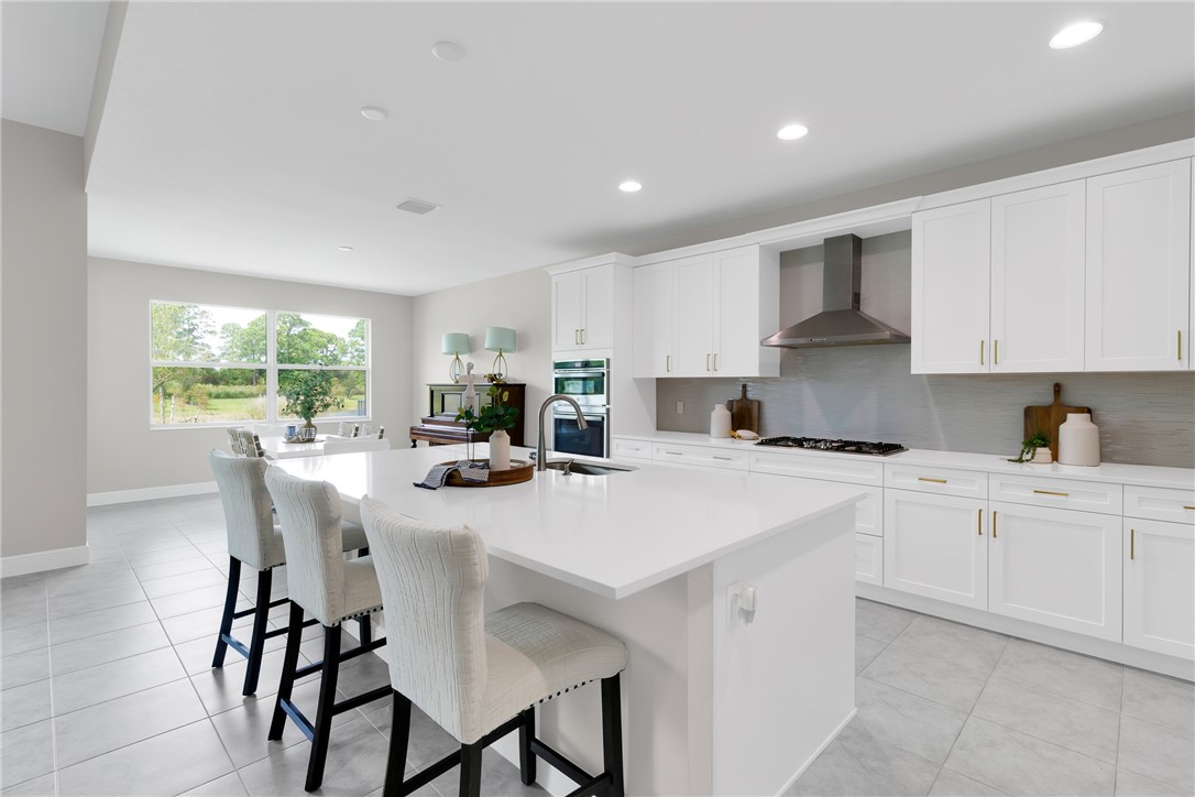 Open-concept kitchen featuring an expansive island with countertop seating, white shaker cabinetry, stainless steel range hood, gas cooktop, and tile flooring