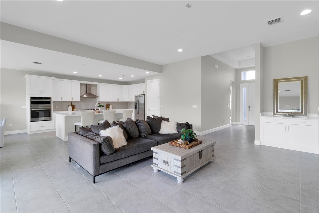 Spacious open-concept living area featuring light gray tile flooring, a modern kitchen with white cabinetry, a built-in double oven, and a stainless steel range hood