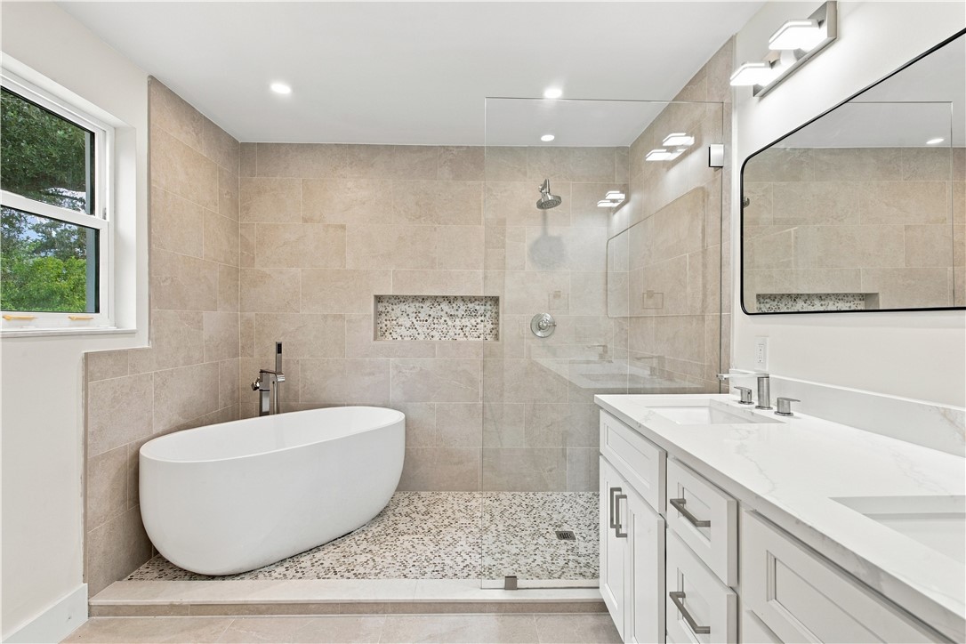 Freestanding soaking tub, walk-in shower with glass enclosure, dual vanity with white cabinetry and countertop, recessed lighting, and tiled walls and flooring