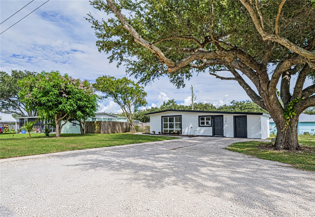 White exterior with dark trim and multiple entry doors, featuring a gravel driveway and a large mature tree