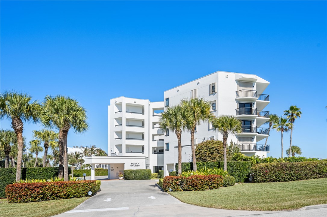 Modern multi-story building featuring multiple balconies, a covered entrance, and extensive landscaping with palm trees and manicured hedges