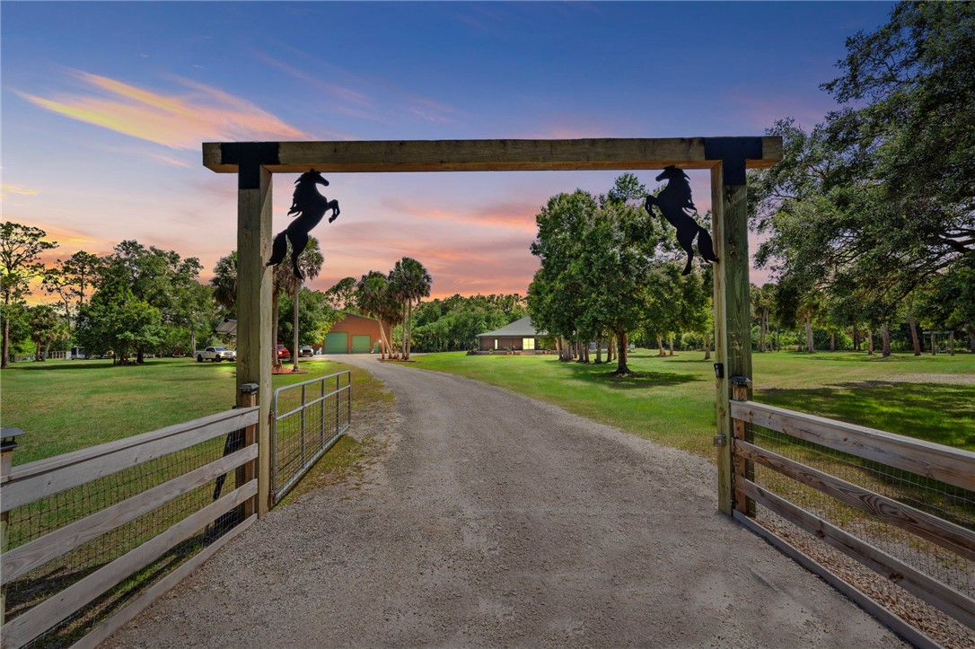 Gravel drive leading to the property, framed by a wooden gate and archway with decorative horse accents, surrounded by expansive green lawns and mature trees