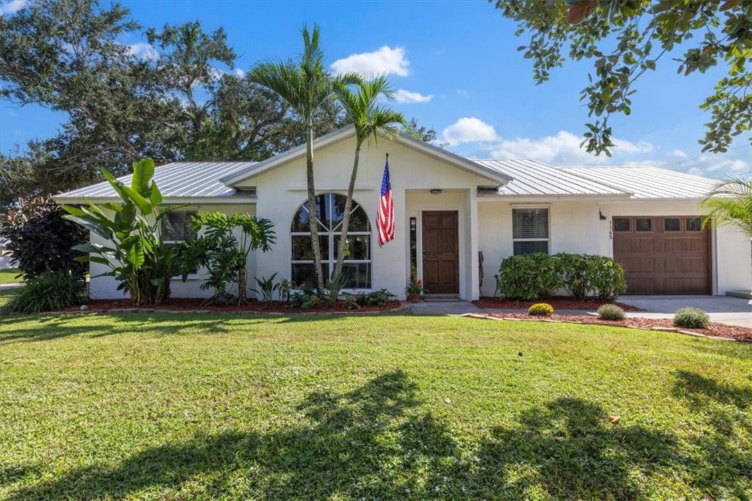 White exterior with a metal roof, an arched window, a paneled front door, a two-car garage with a wood-finish door, and mature landscaping