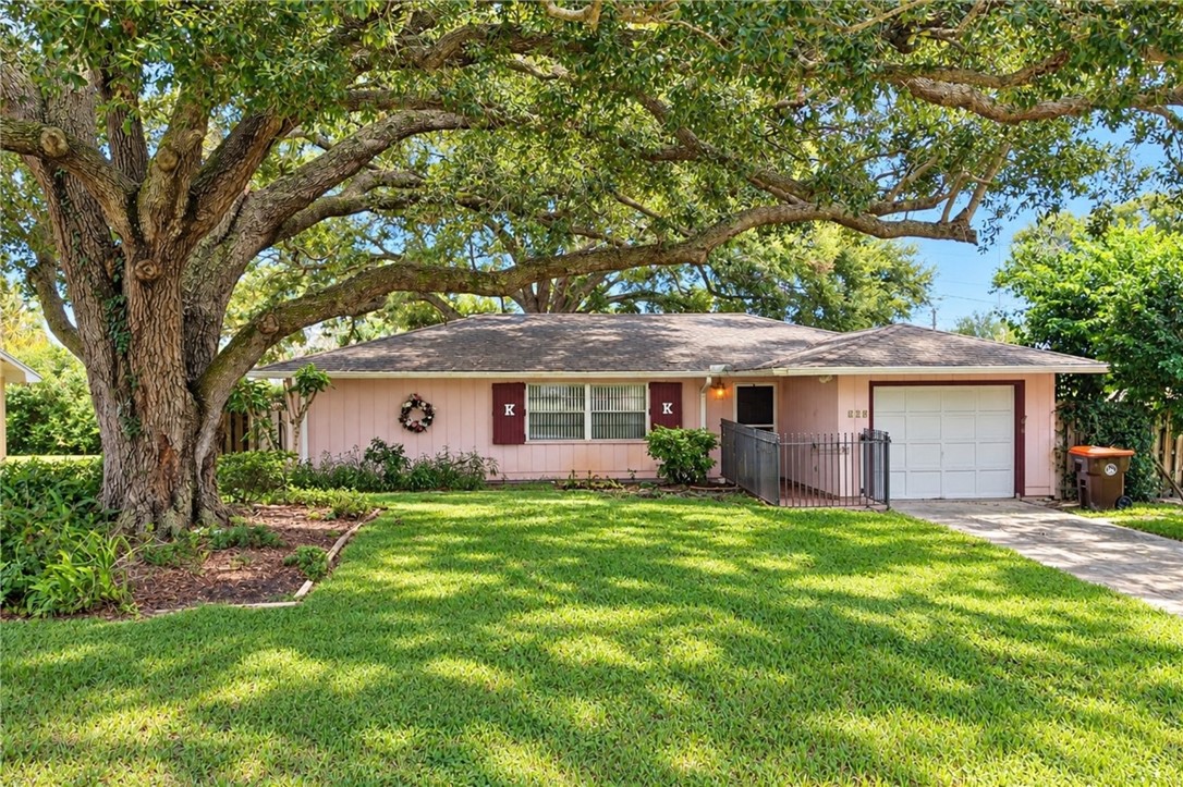 Single-story residence featuring a mature shade tree, a manicured front lawn, a paved driveway, a single-car garage, and a fenced side yard
