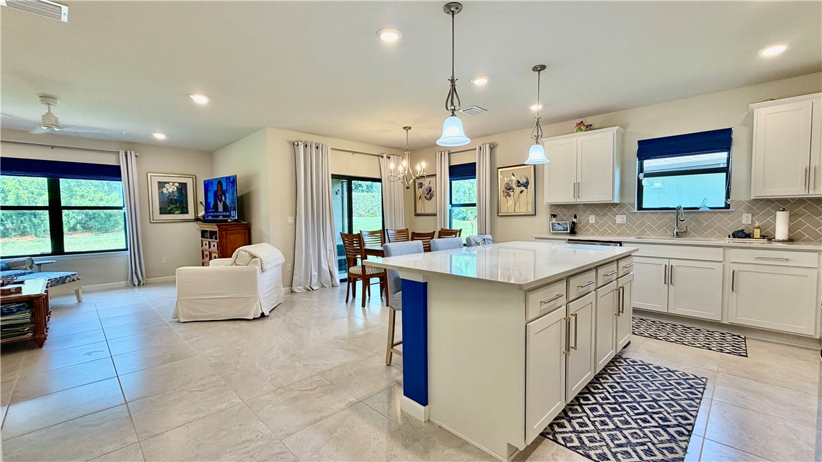 Kitchen featuring a central island with a stone-finish countertop, white cabinetry with gold-tone hardware, pendant lighting, herringbone-patterned tile backsplash, and light-toned floor tiling
