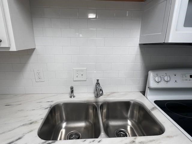 Kitchen featuring a double basin stainless steel sink with a chrome faucet, white subway tile backsplash, light-toned countertop with veining, and white upper cabinetry