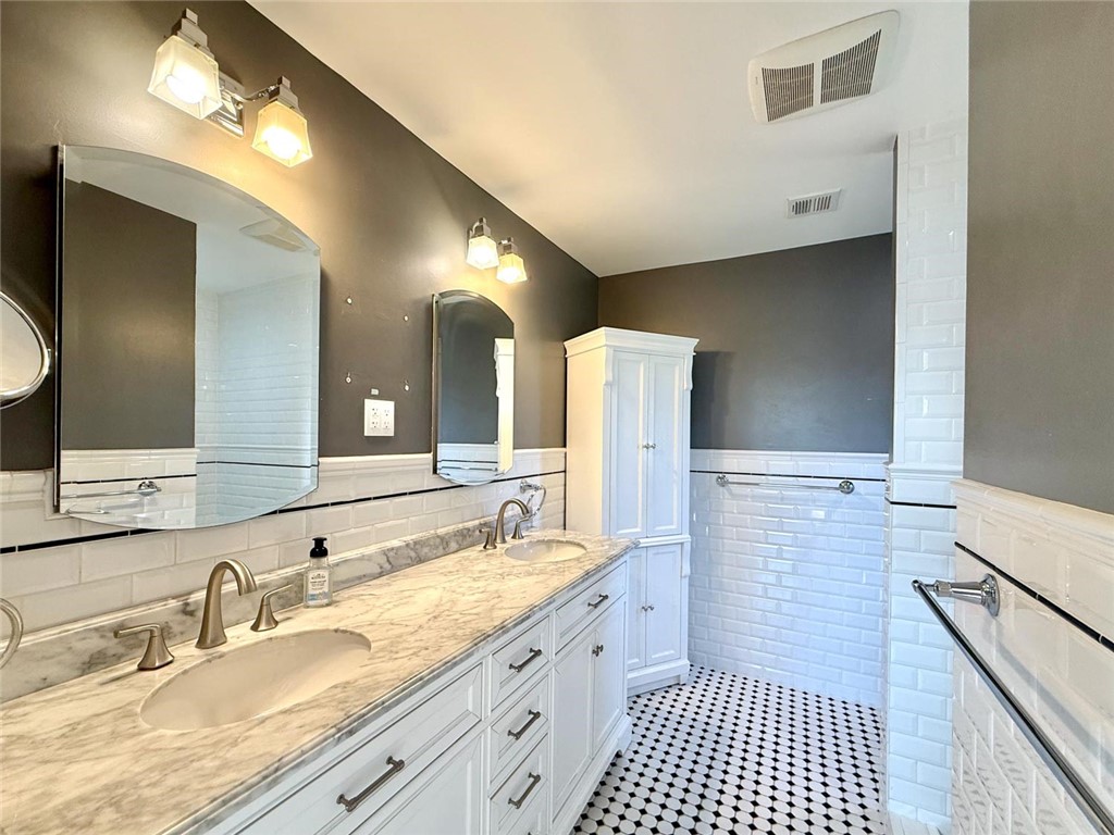 Dual-sink vanity with stone-finish countertop, white cabinetry, ceramic subway tile wainscoting, tall built-in storage cabinet, and monochrome patterned tile flooring