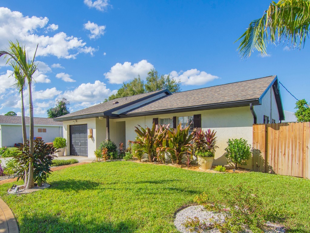 Single-story exterior featuring a light-toned stucco façade, a shingled gable roof, an attached single-car garage, and a wood privacy fence