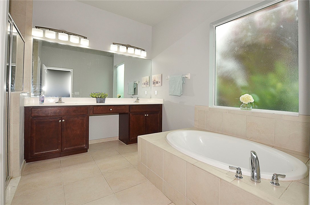 Double vanity with dark wood-finish cabinetry, solid-surface countertop, full-width vanity mirror, large soaking tub with tile surround, and privacy glass window