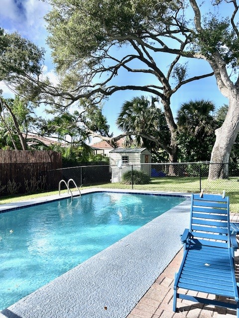 In-ground swimming pool with tile border, paver deck, chain-link fencing, mature trees, and detached storage shed