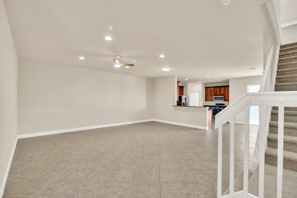Open-concept interior featuring large-format tile flooring, recessed lighting, ceiling fan, carpeted staircase with white balusters, and kitchen with wood cabinetry