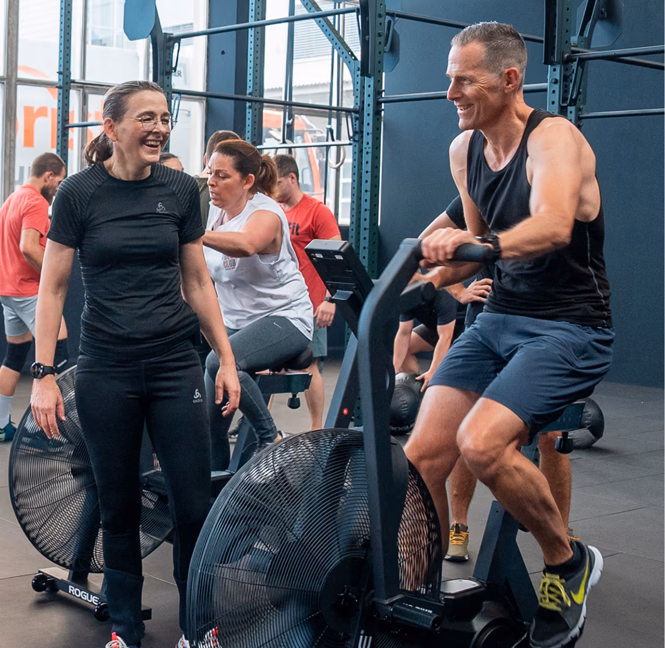 Zwei Erwachsene trainieren in einem Fitnessstudio: Eine Frau in schwarzer Sportkleidung lacht stehend neben einem Mann in schwarzem Tanktop und blauen Shorts, der auf einem Trainingsfahrrad sitzt. Im Hintergrund trainieren weitere Personen an Geräten.