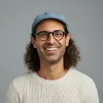 Smiling man with curly hair wearing glasses, a blue cap, and a light sweater against a gray background.