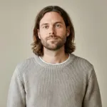 Man with shoulder-length brown hair and beard wearing a gray knit sweater against beige background.