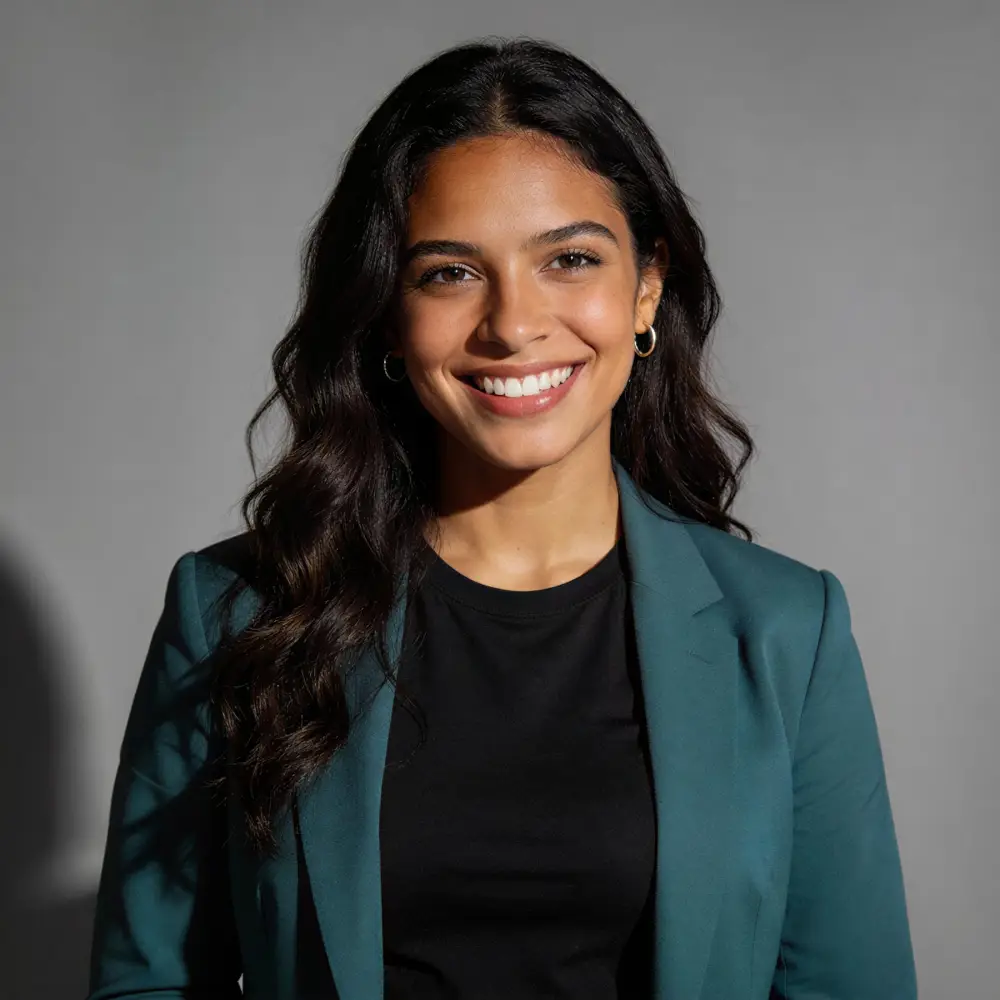 Smiling woman with long dark hair wearing a teal blazer and black shirt against a gray background.