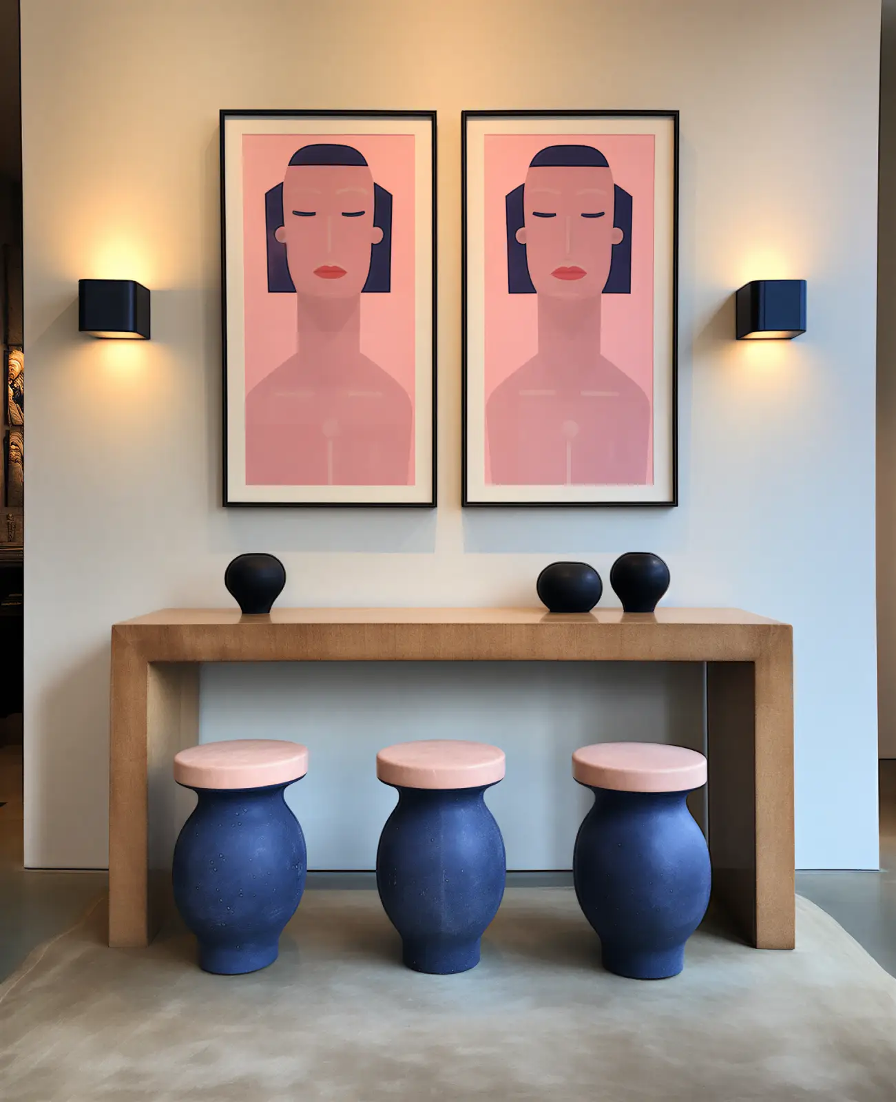 Modern interior featuring a wooden console table with three blue stools topped with pale pink cushions underneath, three black sculptural vases on the table, and two framed minimalist portraits of a face with closed eyes and dark hair on a pink background hanging above, illuminated by two wall sconces on each side.