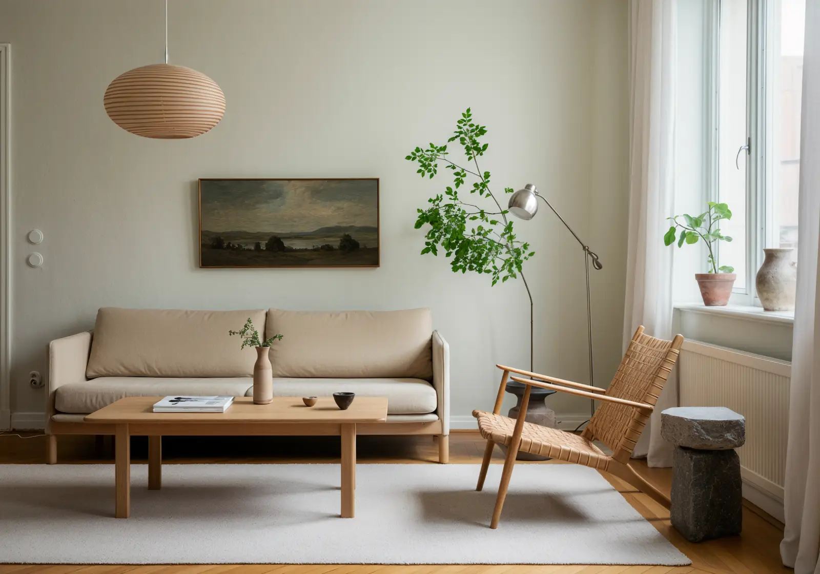 Minimalist living room with a beige sofa, wooden coffee table holding a vase and small decor, a woven chair, a floor lamp with green leafy branches, and two potted plants near a window with sheer curtains.