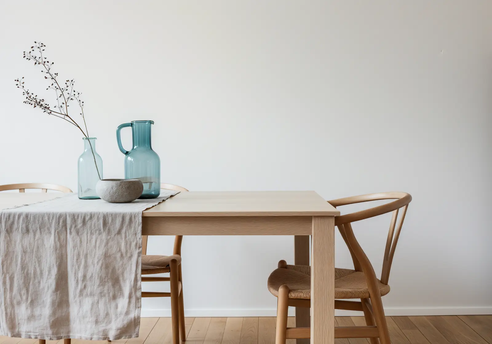 Minimalist dining setup with a light wooden table and two matching chairs on a hardwood floor, featuring a beige linen table runner and decorative blue glass pitcher, glass vase with dried branches, and a small stone bowl on the table.