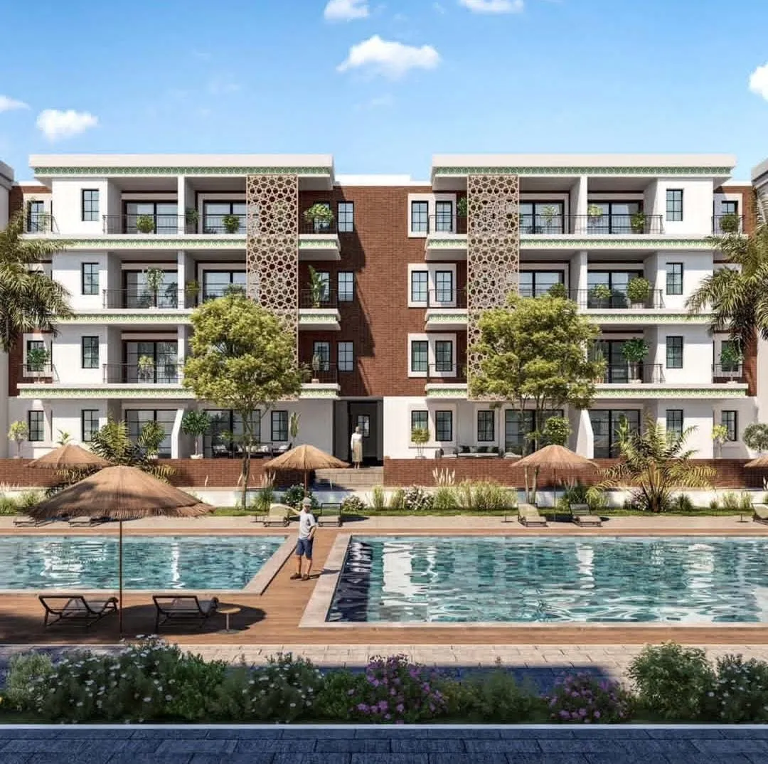 Modern three-story residential building with balconies decorated by plants, facing a swimming pool with wooden deck chairs and thatched umbrellas, under a blue sky with scattered clouds.