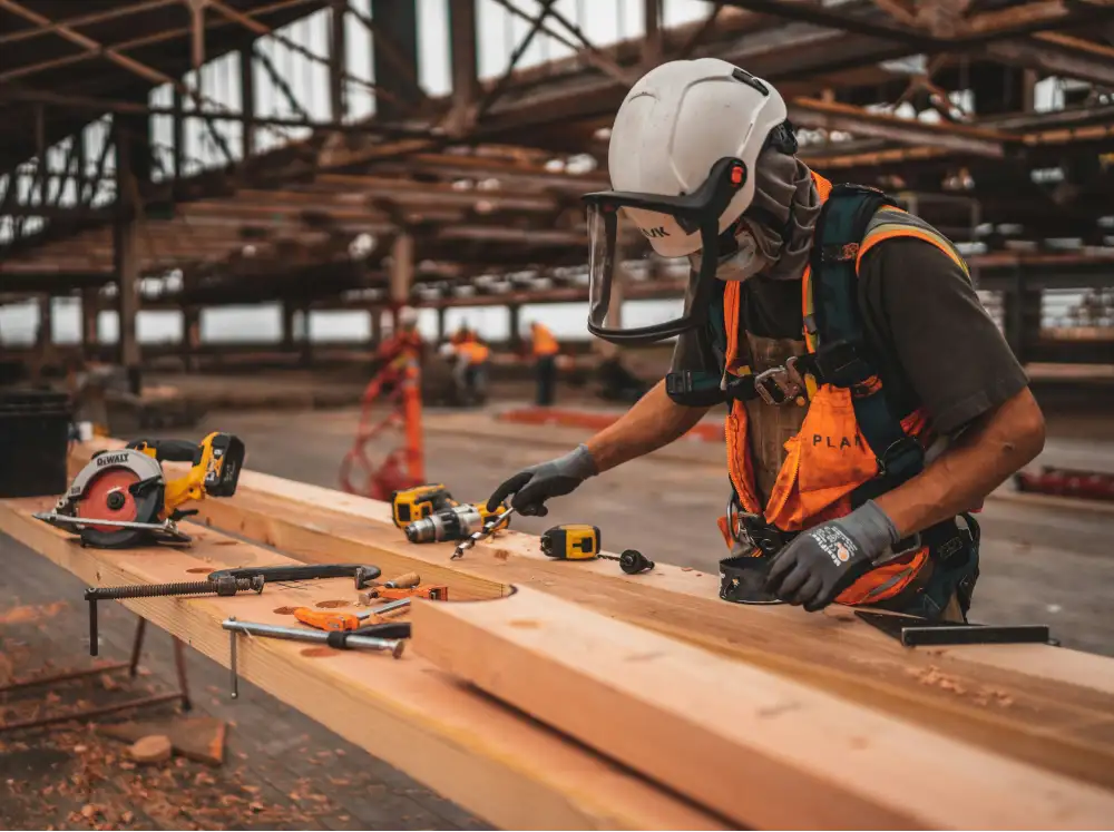 Construction worker wearing a safety helmet with face shield, gloves, and orange safety vest working with wood and power tools in an open warehouse or construction site.