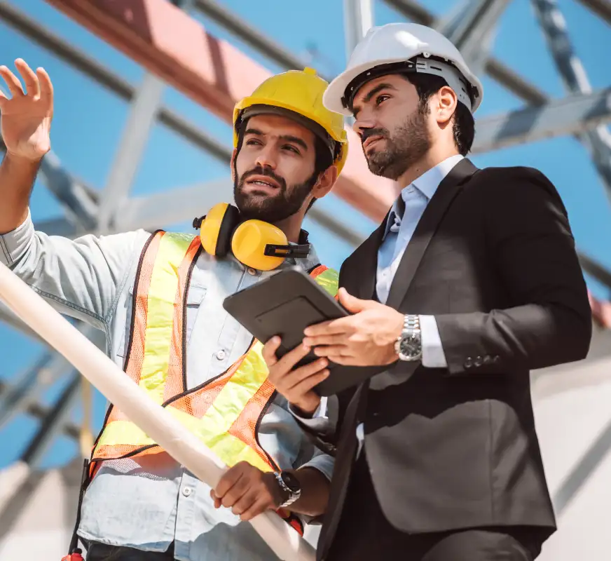 Two men at a construction site, one in a yellow hard hat and safety vest pointing forward holding a pipe, the other in a white hard hat and suit holding a tablet, both looking in the same direction.
