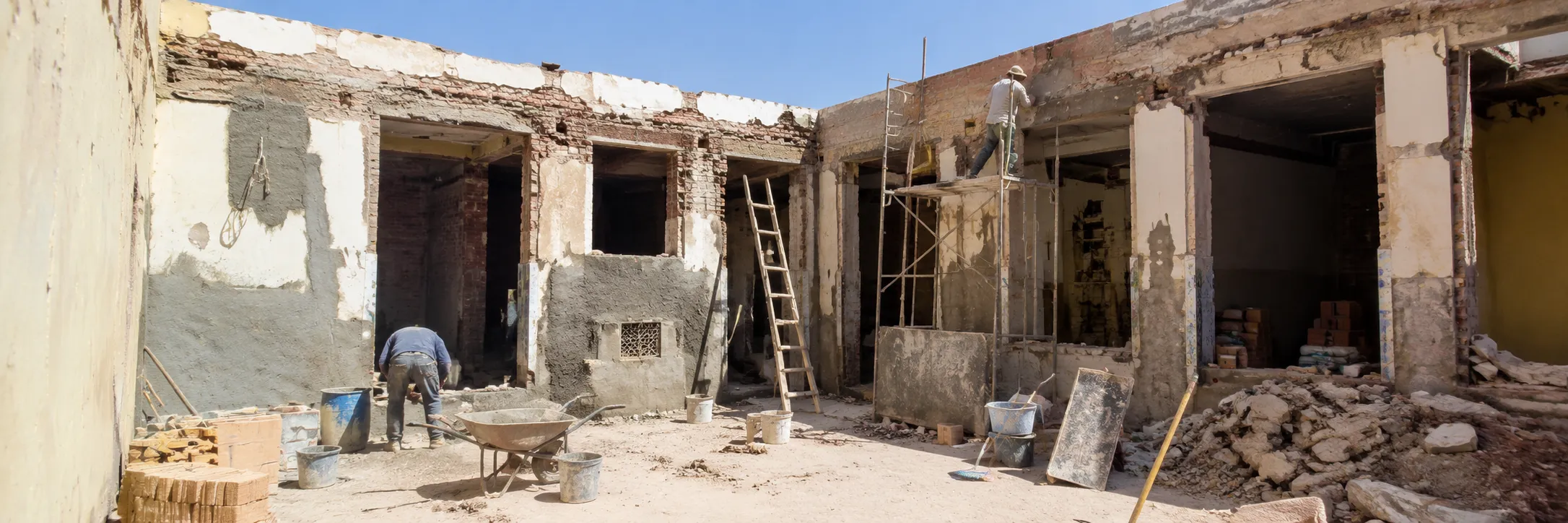 Two construction workers repair and plaster walls of a partially demolished building, with one worker bending down near a wheelbarrow and buckets, and another standing on scaffolding working near the roof under a clear blue sky.