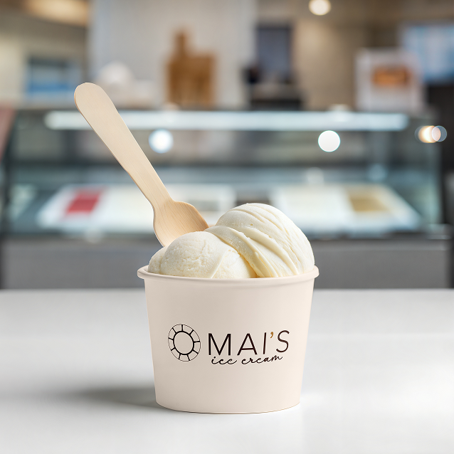 Cup of Mai’s Ice Cream’s vanilla ice cream with two scoops, a wooden spoon, and the Mai’s Ice Cream logo, photographed on a counter inside a softly blurred ice cream shop.