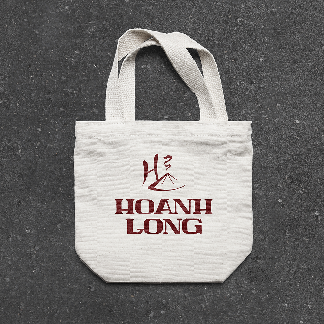 White canvas tote bag featuring the Hoanh Long logo and burgundy typography, displayed against a dark textured background.