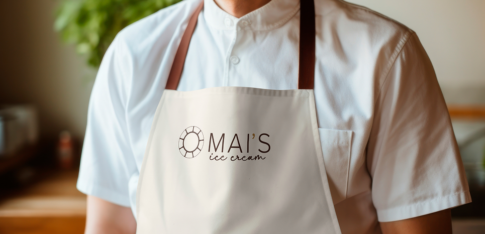 Ice cream shop employee wearing a white apron featuring the Mai’s Ice Cream logo in a warm, softly blurred interior.