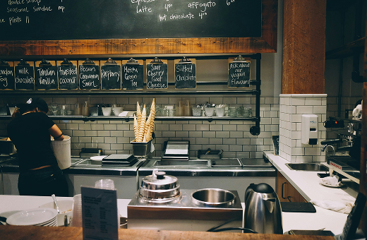 Interior of an artisanal ice cream shop with a tiled counter, handwritten flavor boards, waffle cones, and staff working behind the counter.