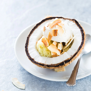 Coconut ice cream served inside a halved coconut shell, topped with toasted coconut flakes and presented on a white plate.
