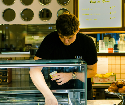 Ice cream shop employee serving from a display case, with menu boards, cups, and pastries visible in the background.