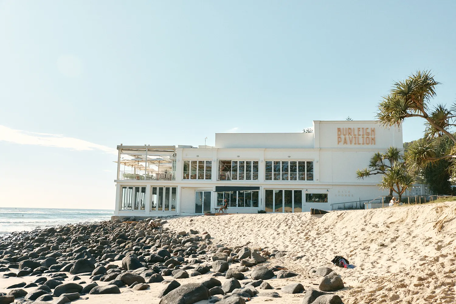 Burleigh Pavilion building seen from the beach, with rocks and palms in the foreground