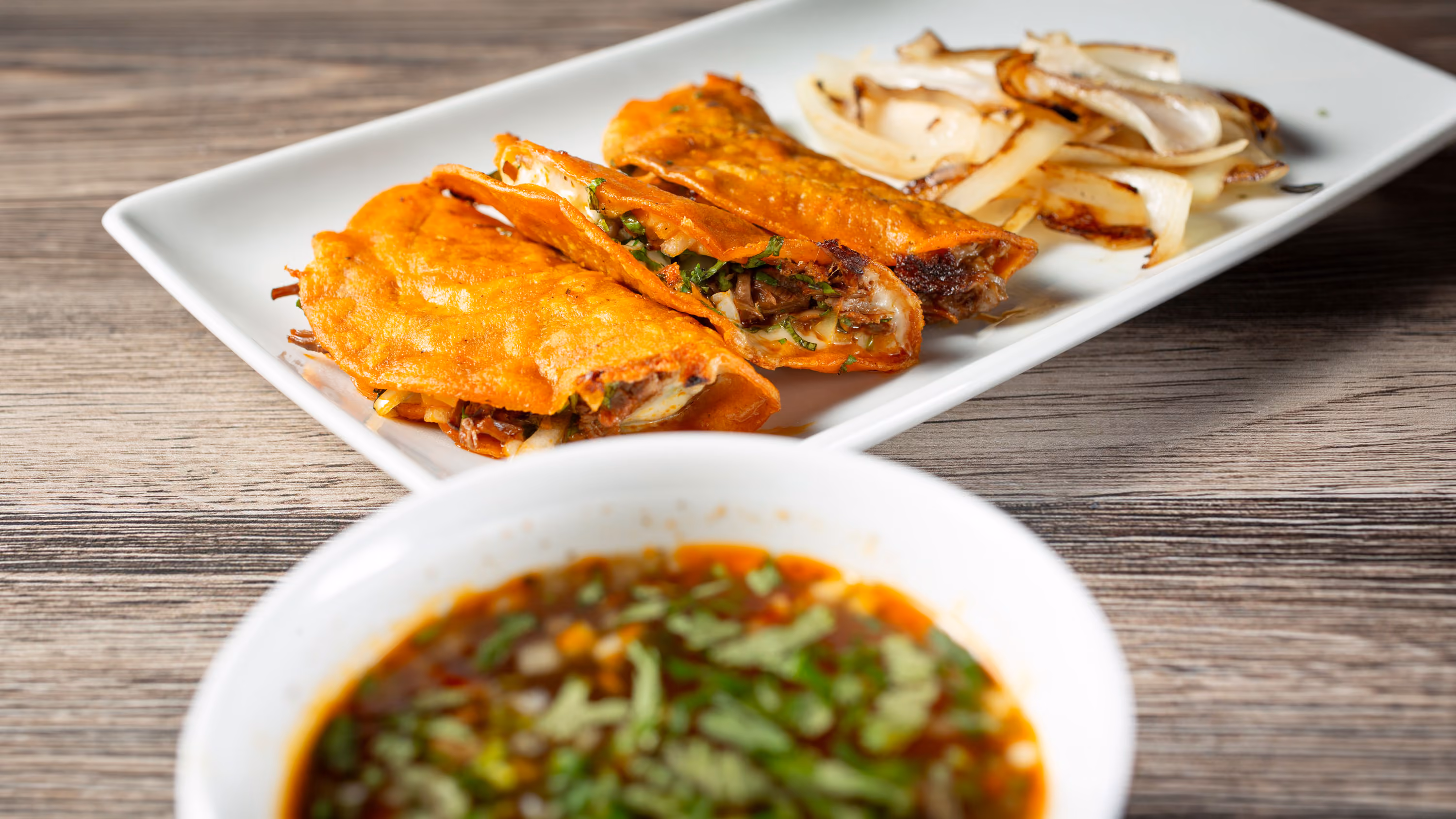 Three crispy birria tacos filled with meat and herbs on a plate with grilled onions and Consome.