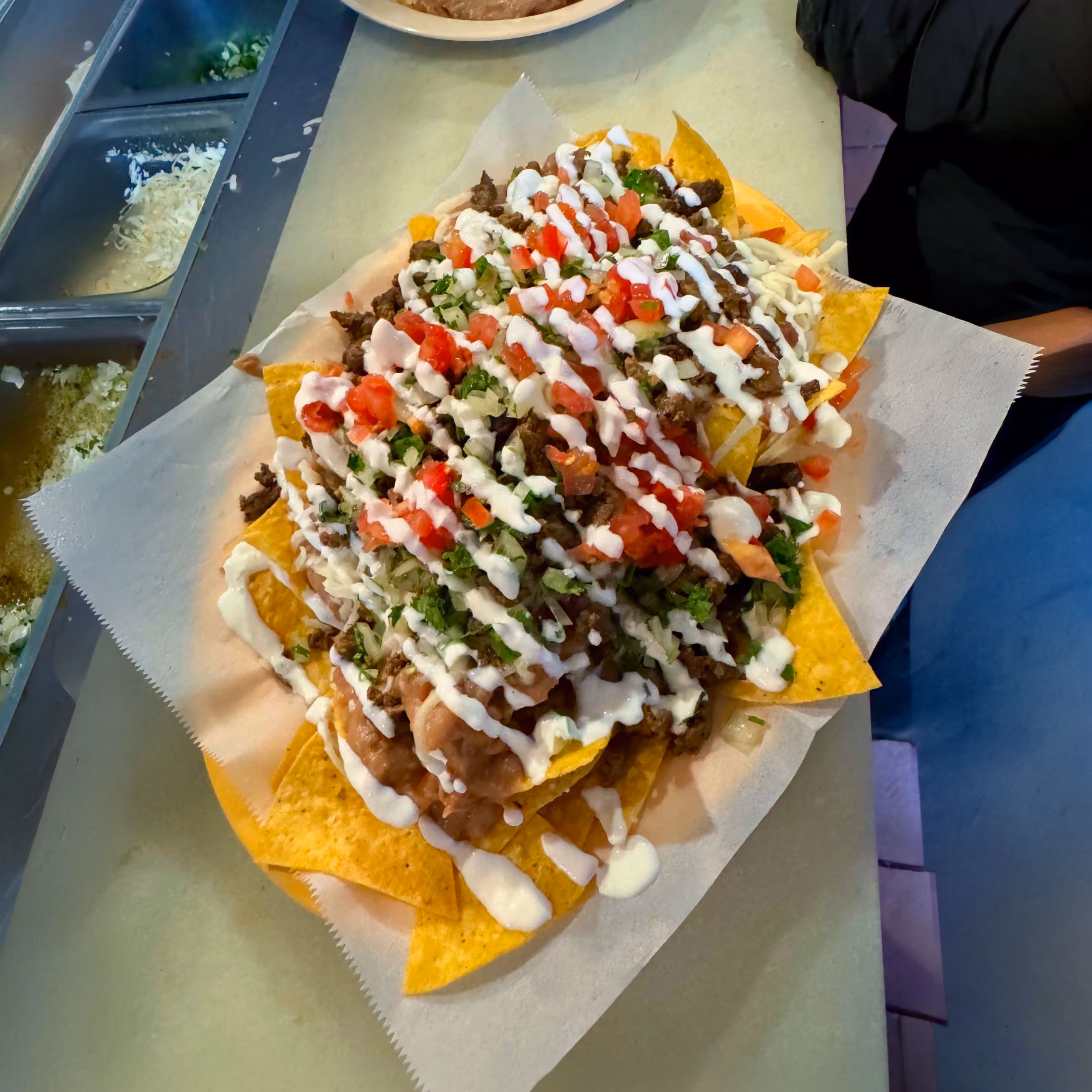 Plate of nachos topped with ground beef, refried beans, tomatoes, cilantro, onions, and drizzled with white sauce.