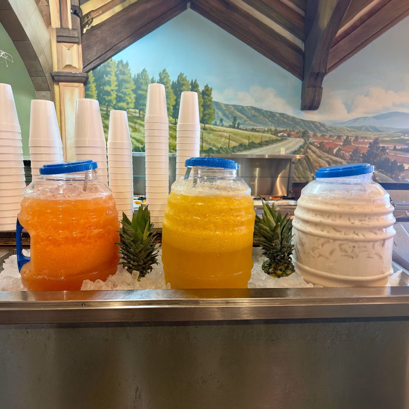 Three large containers with orange, yellow, and white drinks on ice, with stacks of white cups behind.