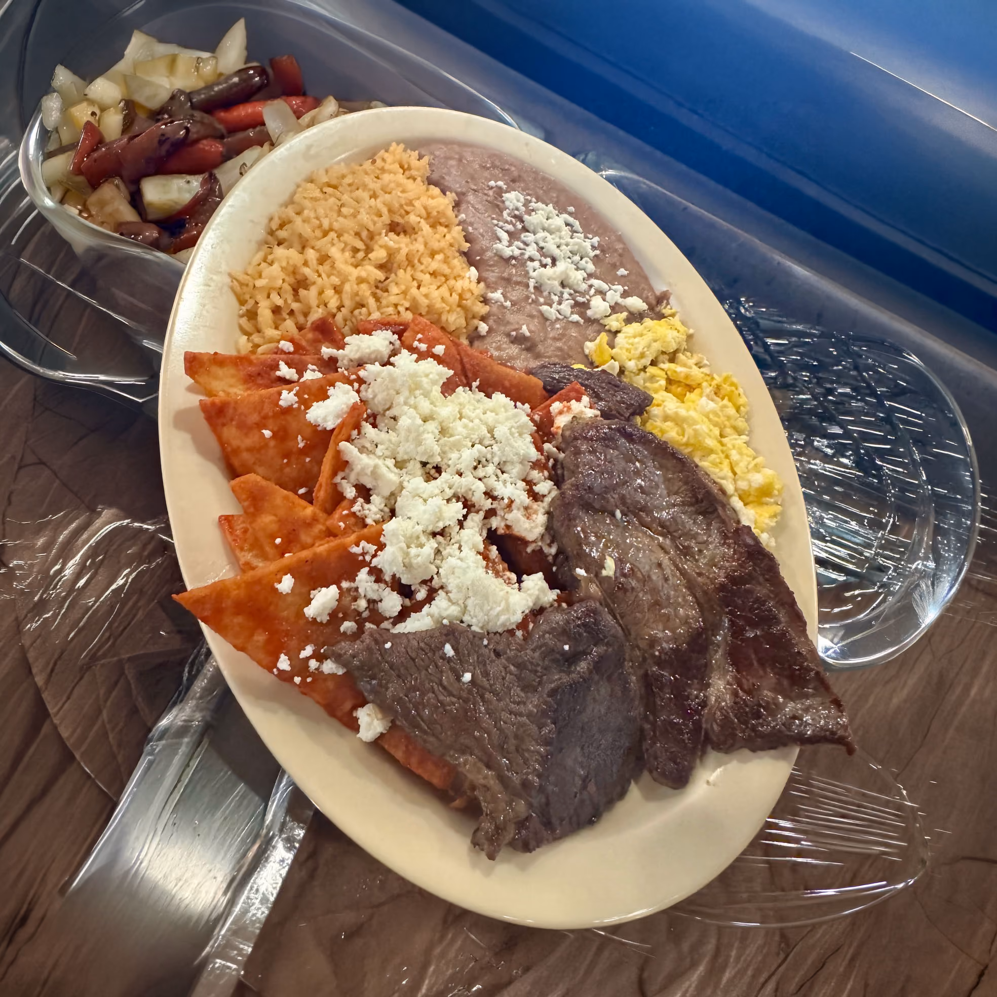 Plate with chilaquiles topped with cheese, beef slices, scrambled eggs, rice, and refried beans with cheese.