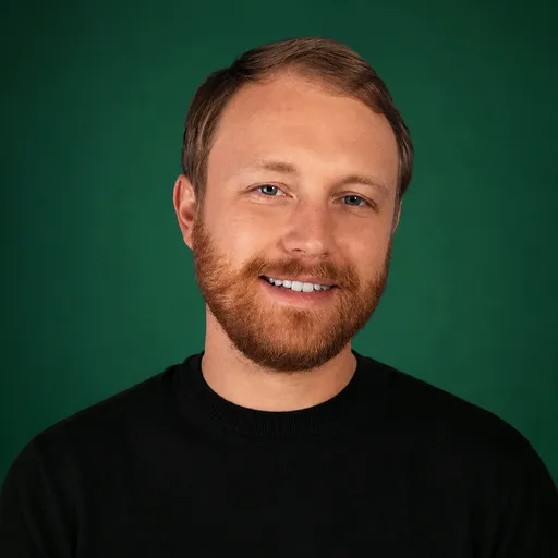 Portrait of a young man with light skin, red hair and beard, wearing a black shirt, smiling against a dark green background.