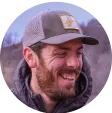 Smiling man with a beard wearing a baseball cap and jacket, outdoors with blurred background.